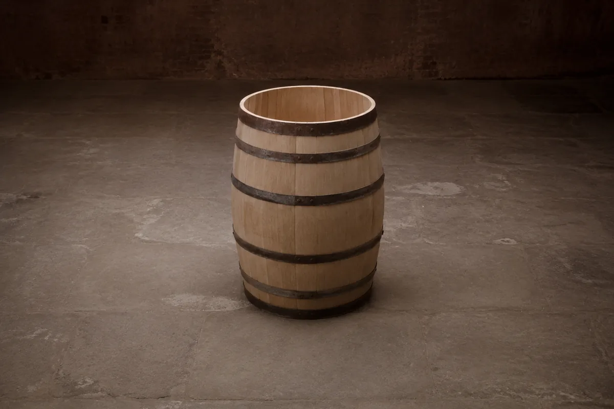 A single oak barrel standing upright and alone on a bare stone floor, its open head towards the camera and its pale uncharred inner staves visible, with nothing else in the room.