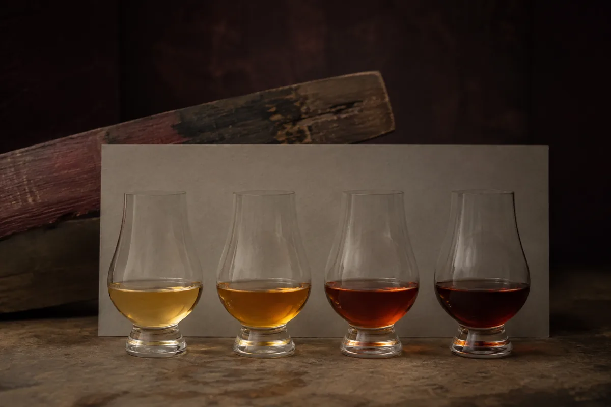 Four identical whisky samples from pale gold through reddish amber to deep brown beside one wine-stained oak stave.