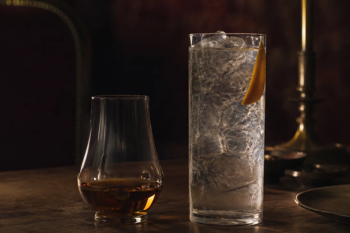 A modest neat rum sample beside a tall rum highball containing abundant clear mixer, ice, and a restrained citrus peel.