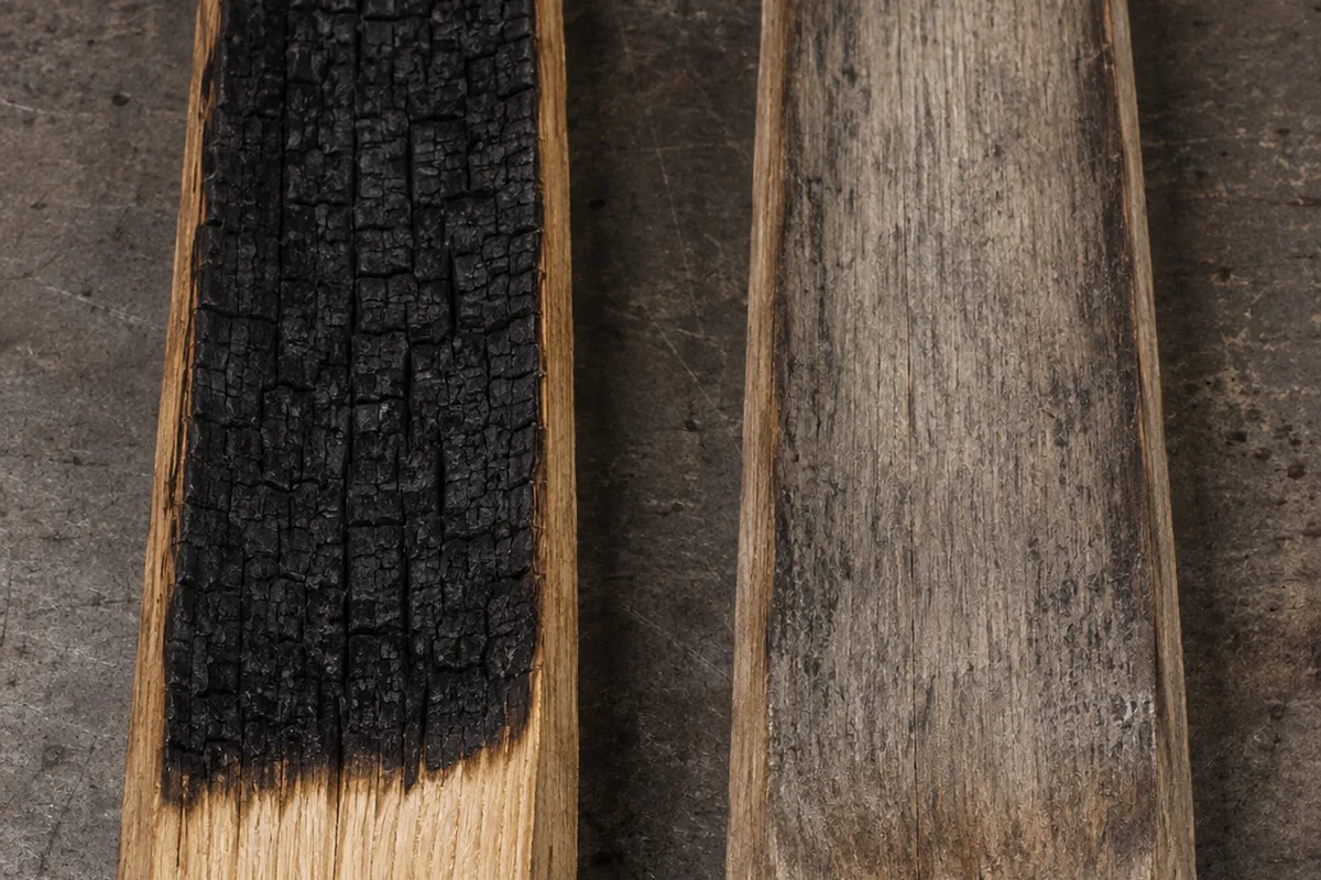 Two oak staves side by side on a workbench, the left with deep black char over fresh pale oak and the right grey, spent and worn smooth